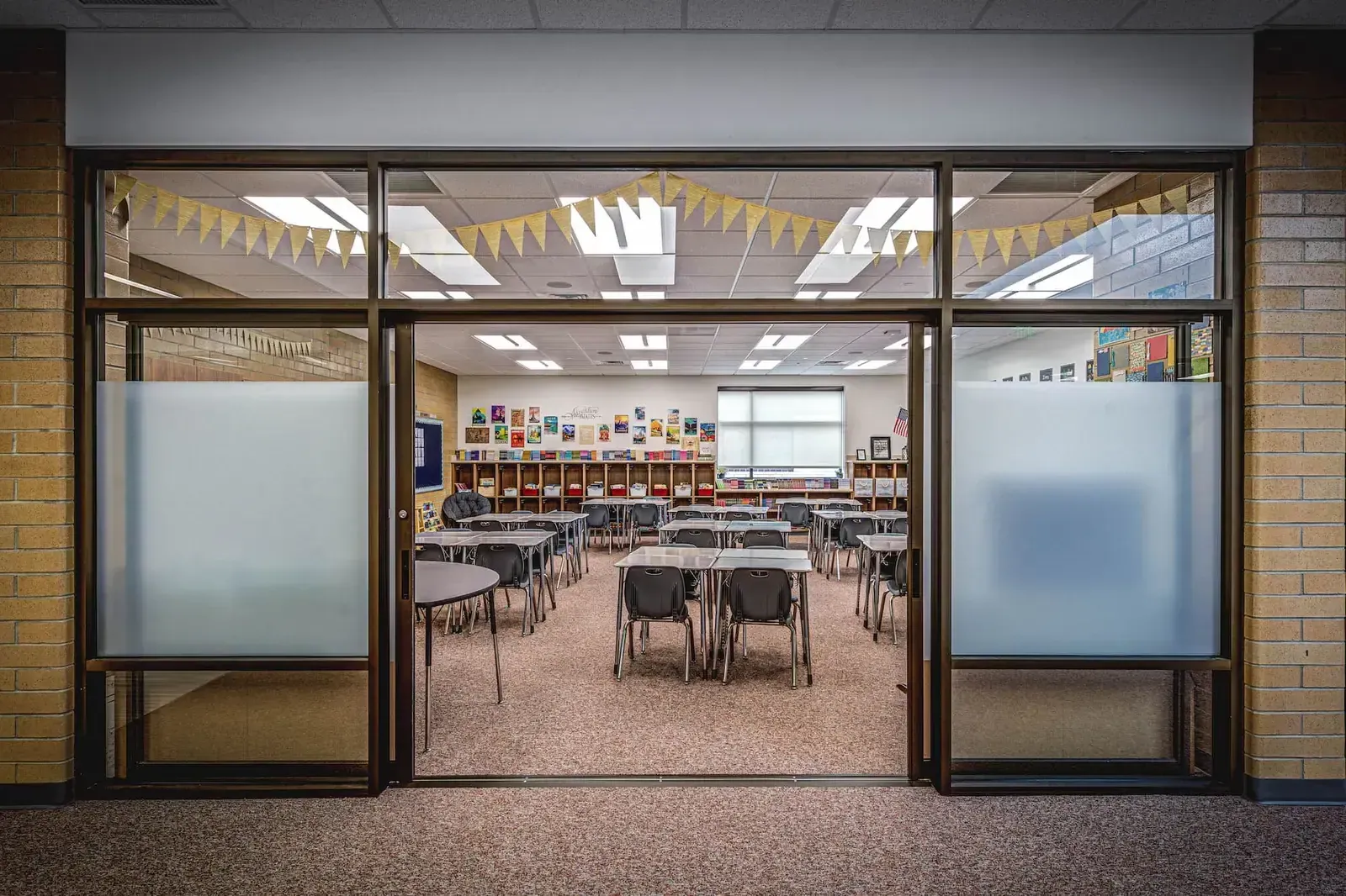 Classroom seen through a glass doorway with frosted panels, with desks grouped inside and a pennant banner overhead.
