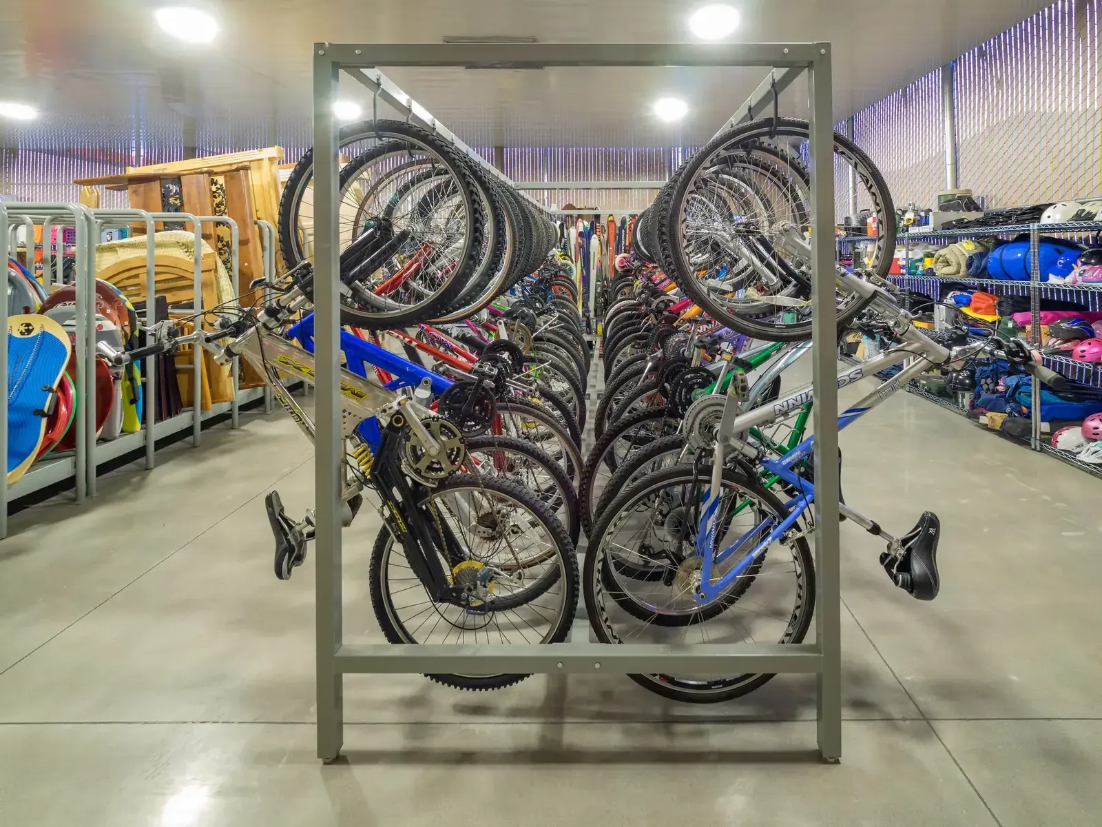 Rows of bicycles hung on a tall metal rack in a large room with shelves of sports gear behind.