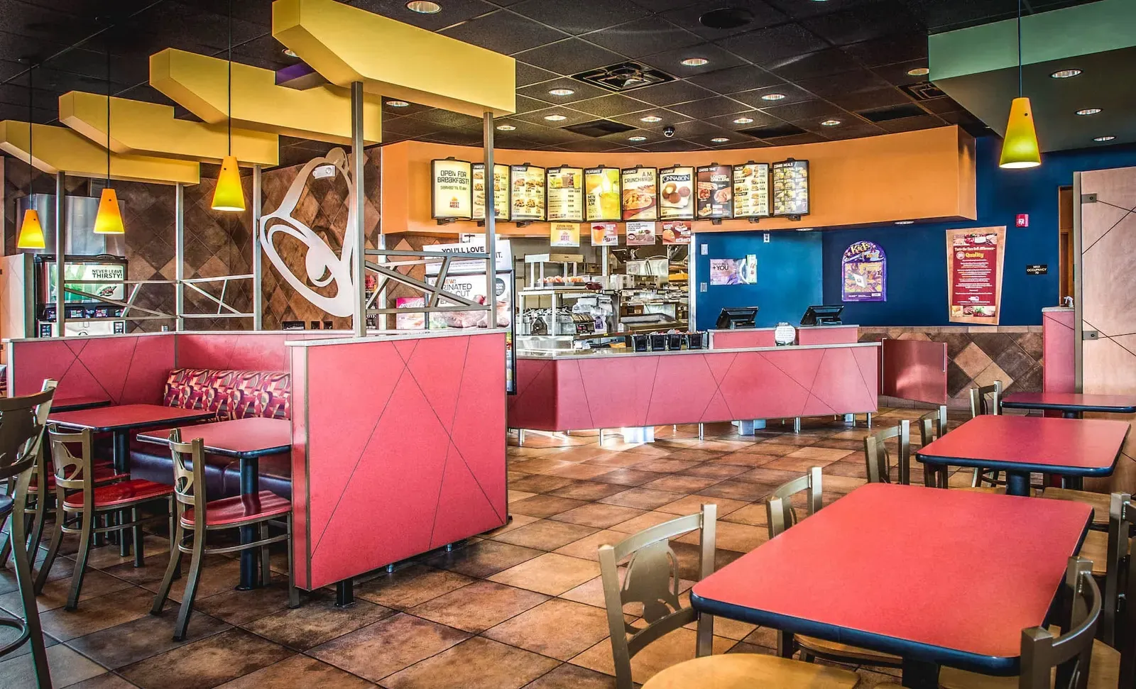 Dining room with red booths and tables, hanging yellow lights and a menu board above the counter.