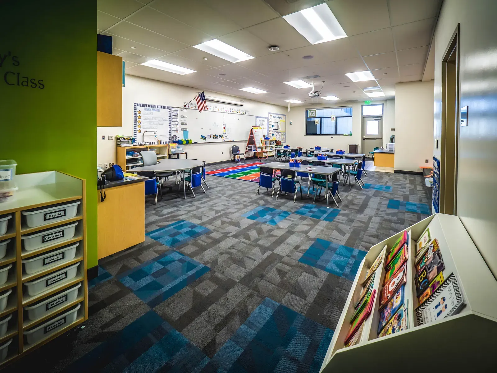 Classroom with small group tables, storage cubbies, a flag and blue-gray patterned carpet