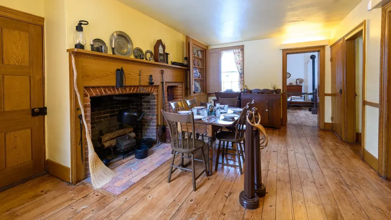 Dining room with a brick fireplace and wood mantel, a set table with spindle-back chairs and wide pine floorboards.