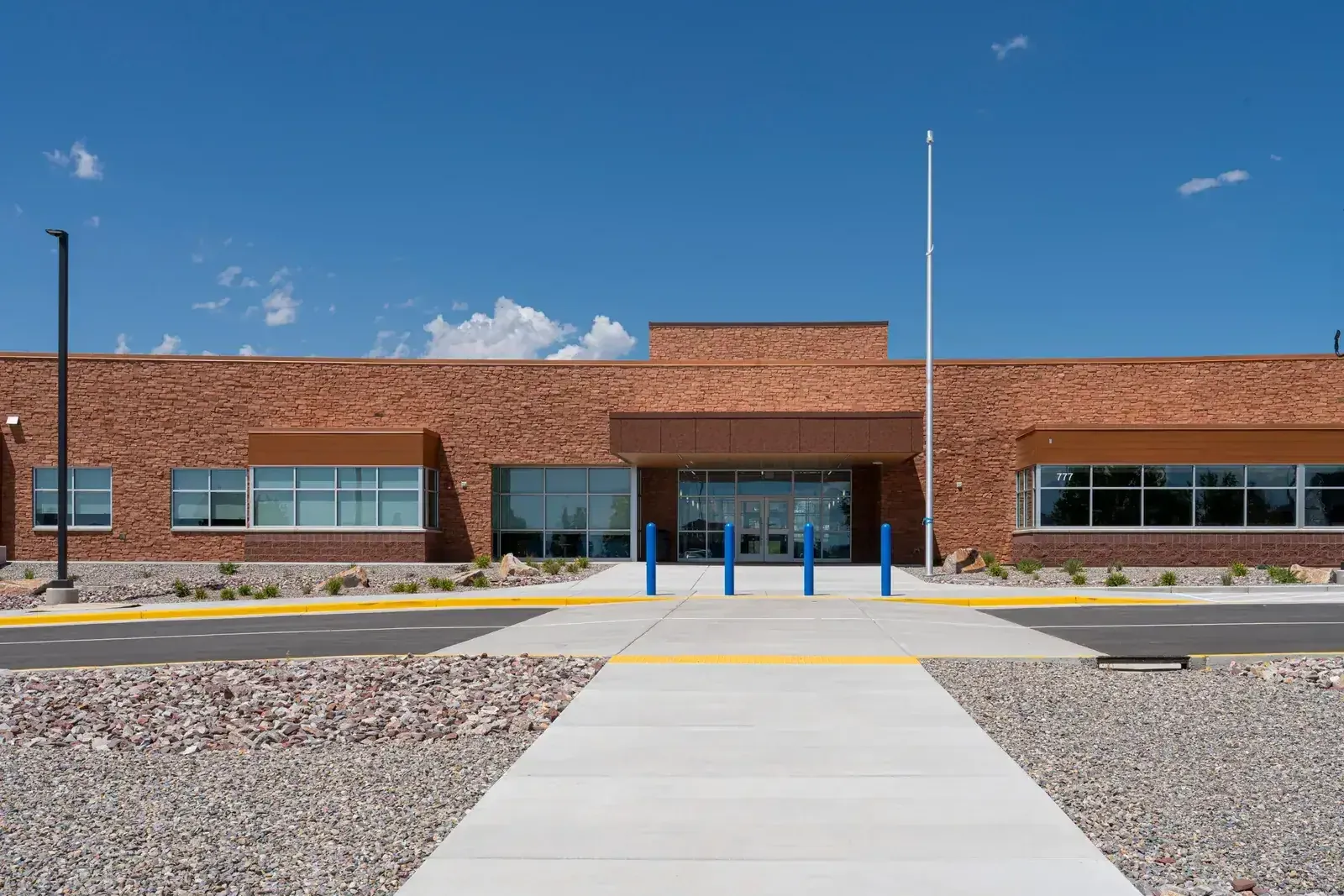 Front entrance of the school with a brick facade, a covered entry, blue bollards and a concrete walkway.