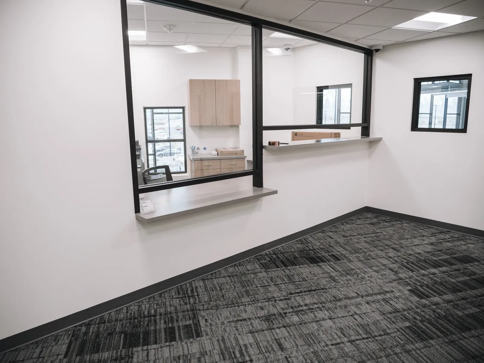 Service window with a black metal frame and a counter looking into an office with wood cabinets.