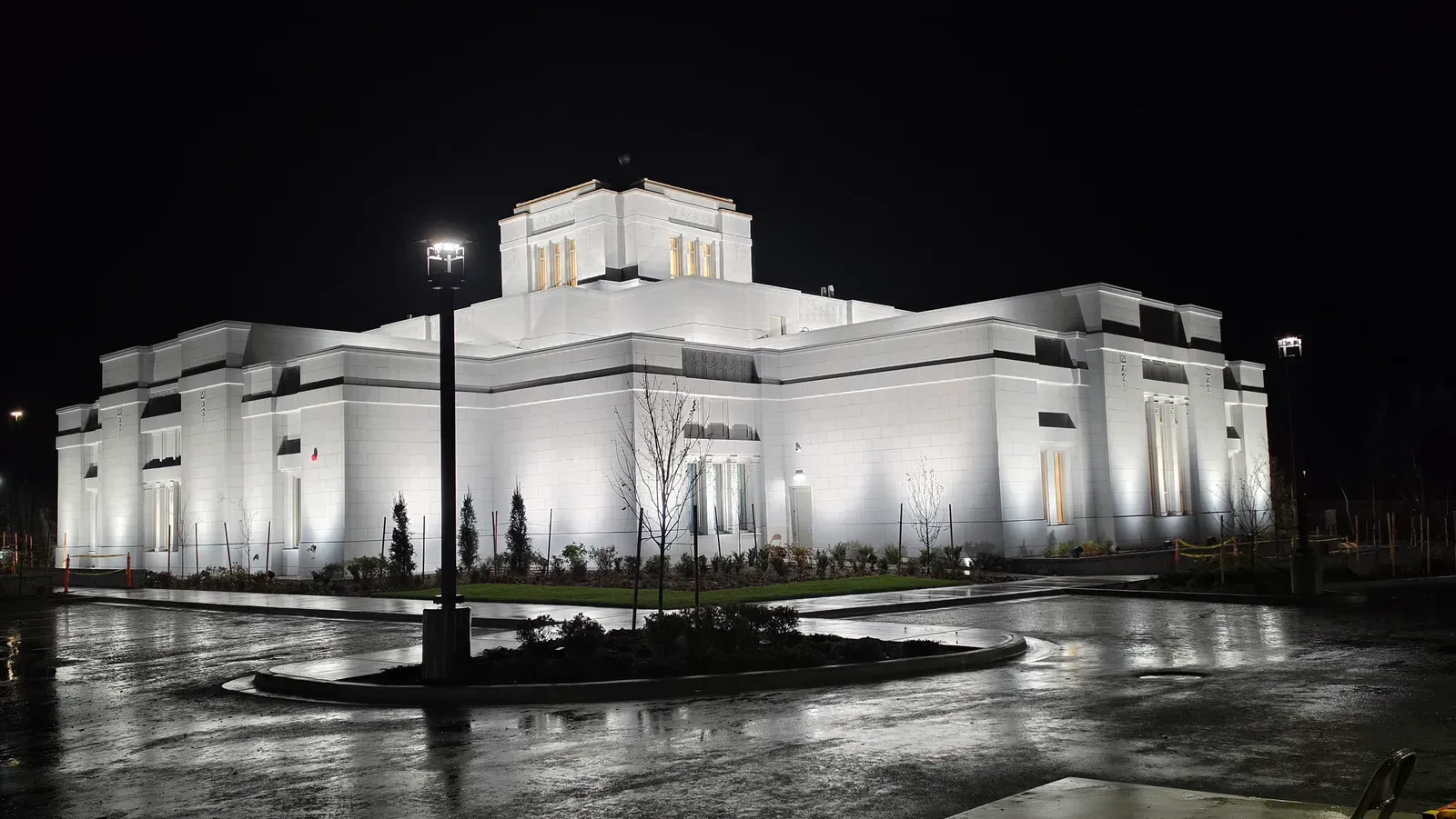 The temple lit by floodlights at night, with its pale stone walls reflected on wet pavement in the foreground.