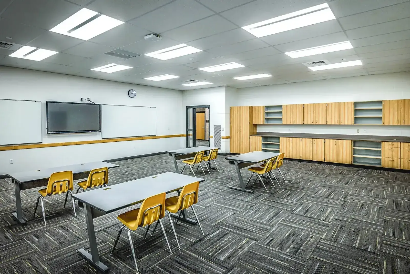 Classroom with gray desks, yellow chairs, a wall-mounted screen and wood cabinets.