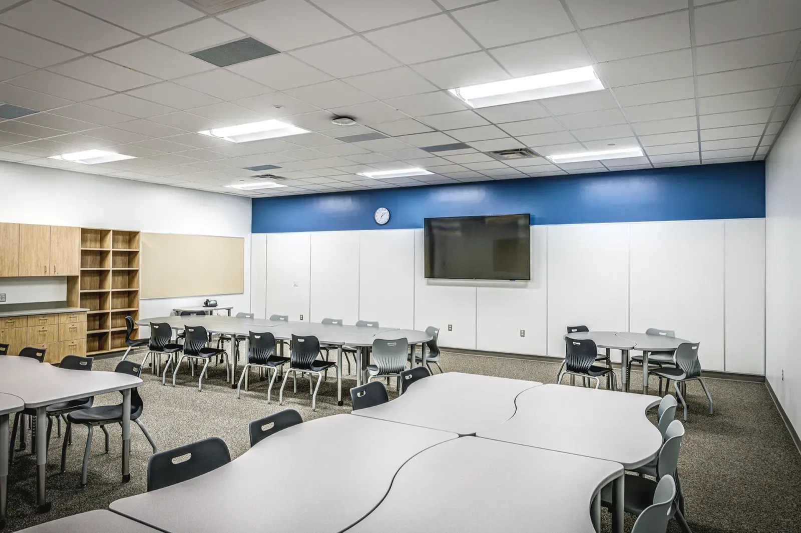 Classroom with curved white tables, gray chairs and a blue wall behind a wall-mounted screen.