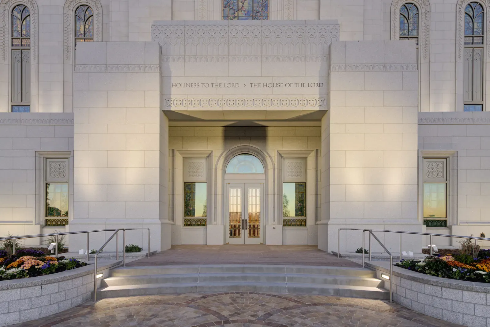 Temple front doors under a stone portico with an inscription above, lit at dusk.