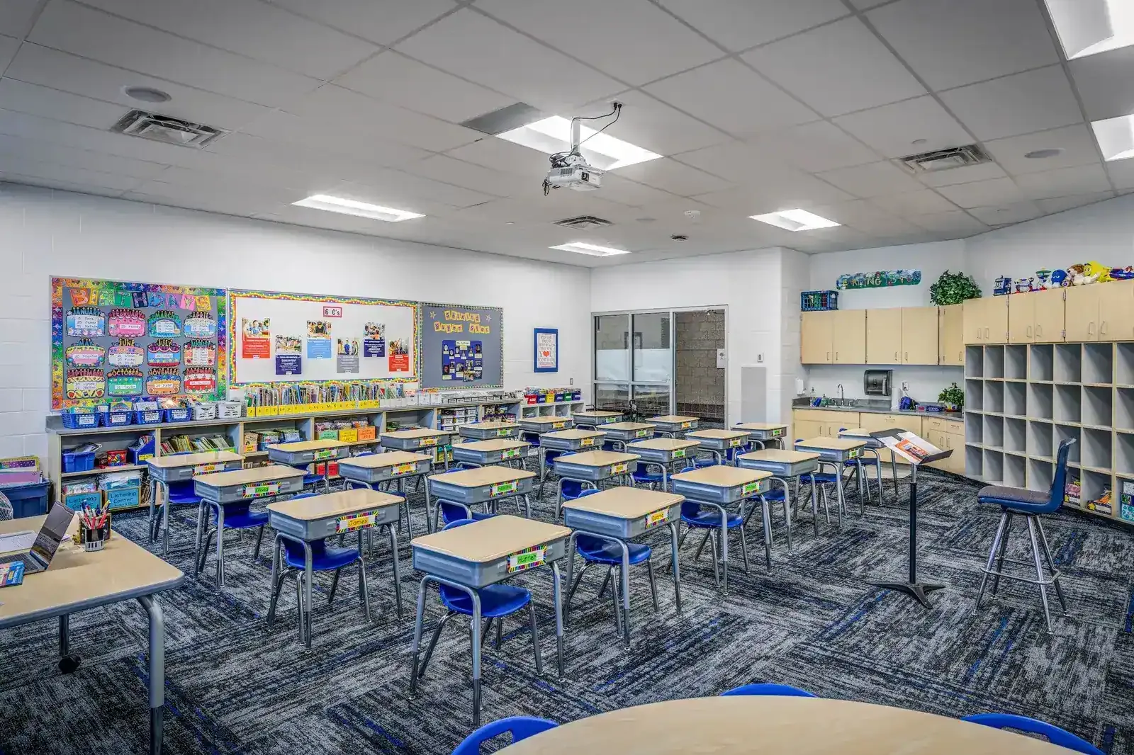 Classroom with student desks, a ceiling projector, shelves and carpeted floor.