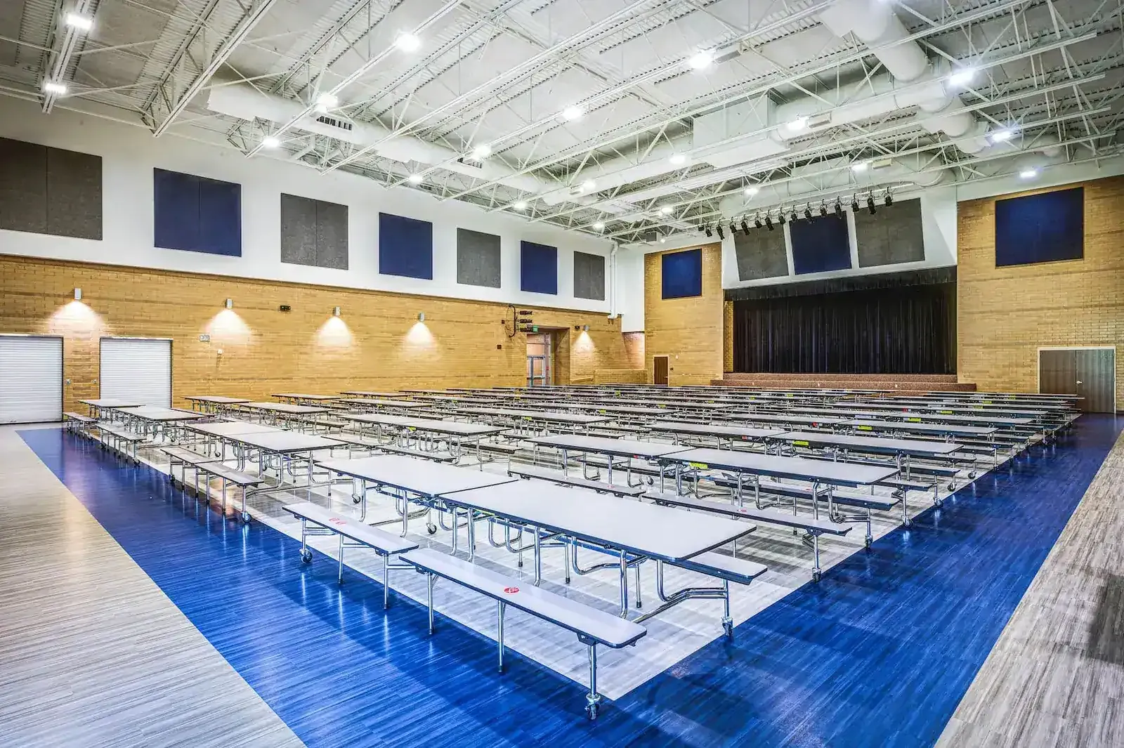 Cafeteria with long folding tables on a blue floor, a curtained stage and acoustic wall panels.