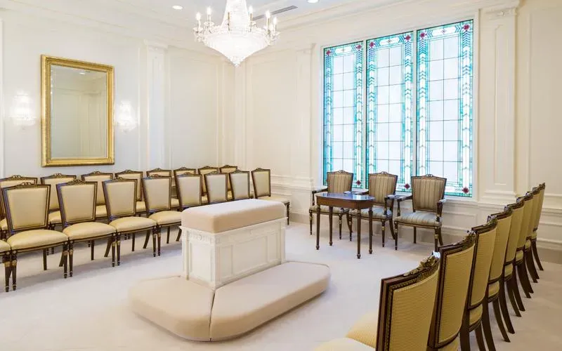 Sealing room with a central cushioned altar, rows of striped upholstered chairs, a gold-framed mirror and a teal stained-glass window