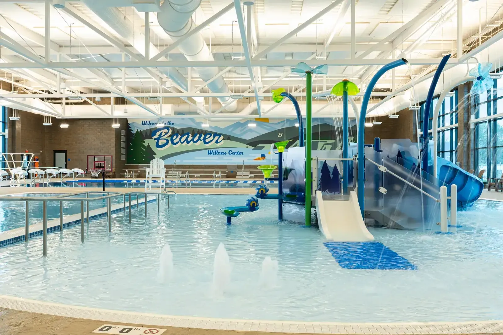Activity pool with water jets, a slide and sculpted play features, and a Welcome to the Beaver Wellness Center mural behind it.