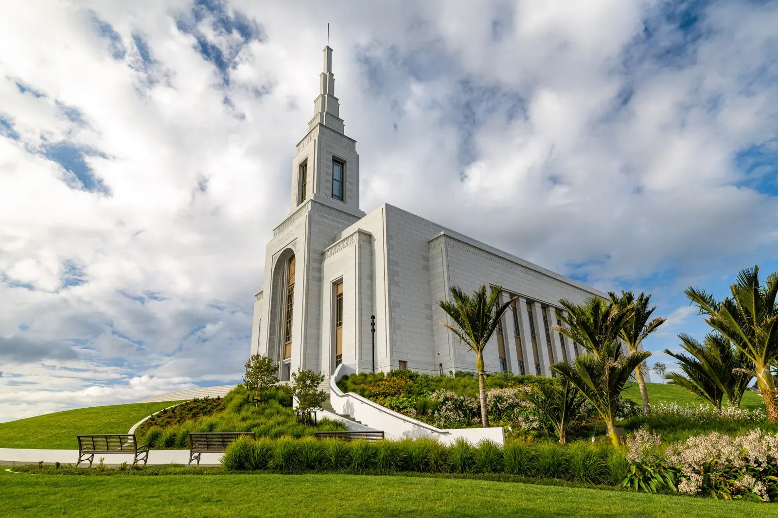 The temple and its spire seen from a low angle under a cloudy blue sky, with palm trees and flowering shrubs in the foreground