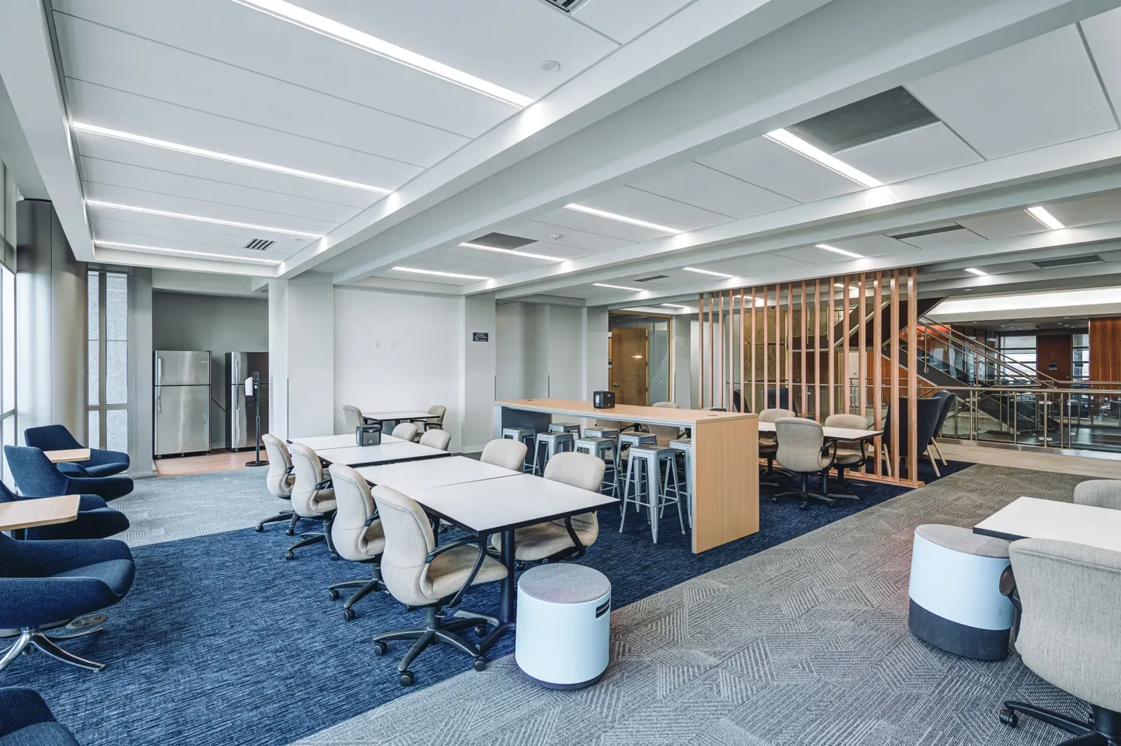 Student commons with tables, chairs and a high counter on blue carpet, with a wood-slat partition and a glass-railed stair behind.