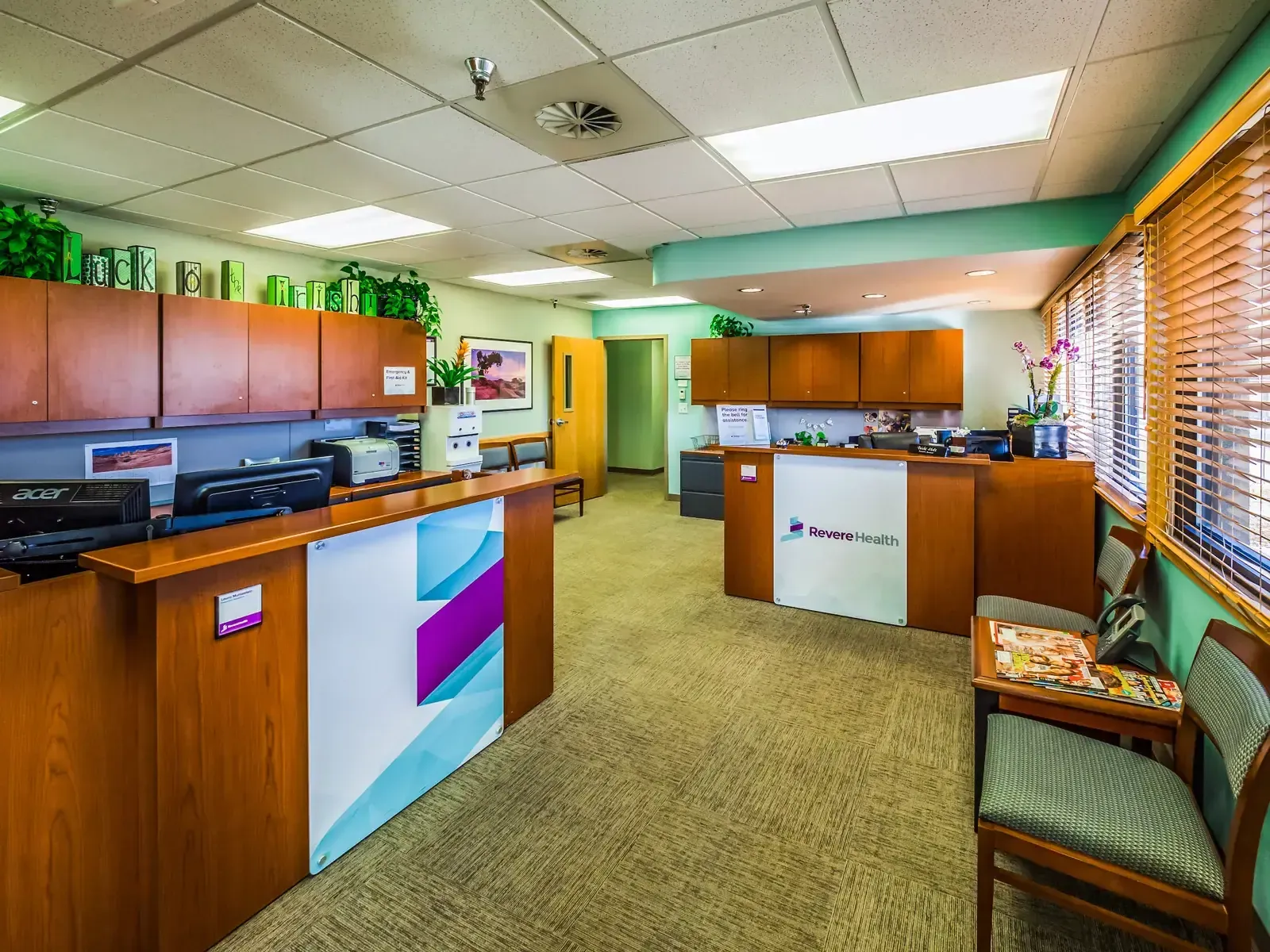 Reception counters with wood cabinetry and Revere Health signage, and a waiting chair beside the window blinds.