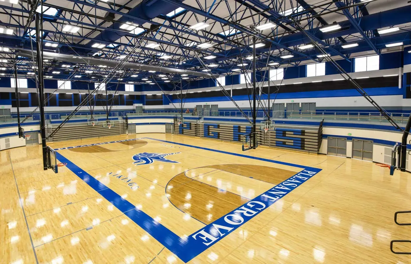 Gymnasium with a hardwood court marked Pleasant Grove, blue steel trusses and bleachers along the sides.