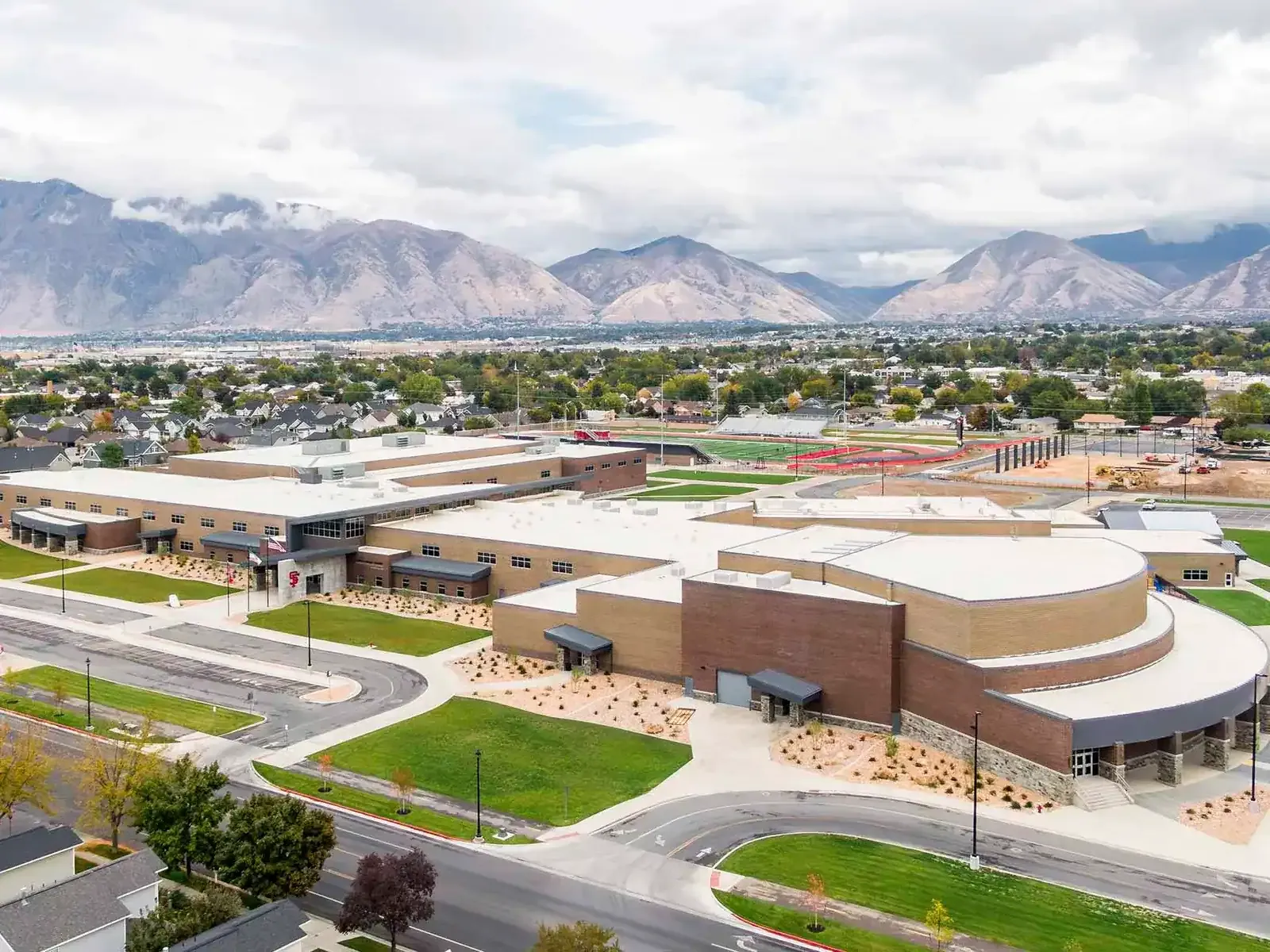 Aerial view of the school campus with its rounded auditorium wing and athletic fields, with mountains behind.