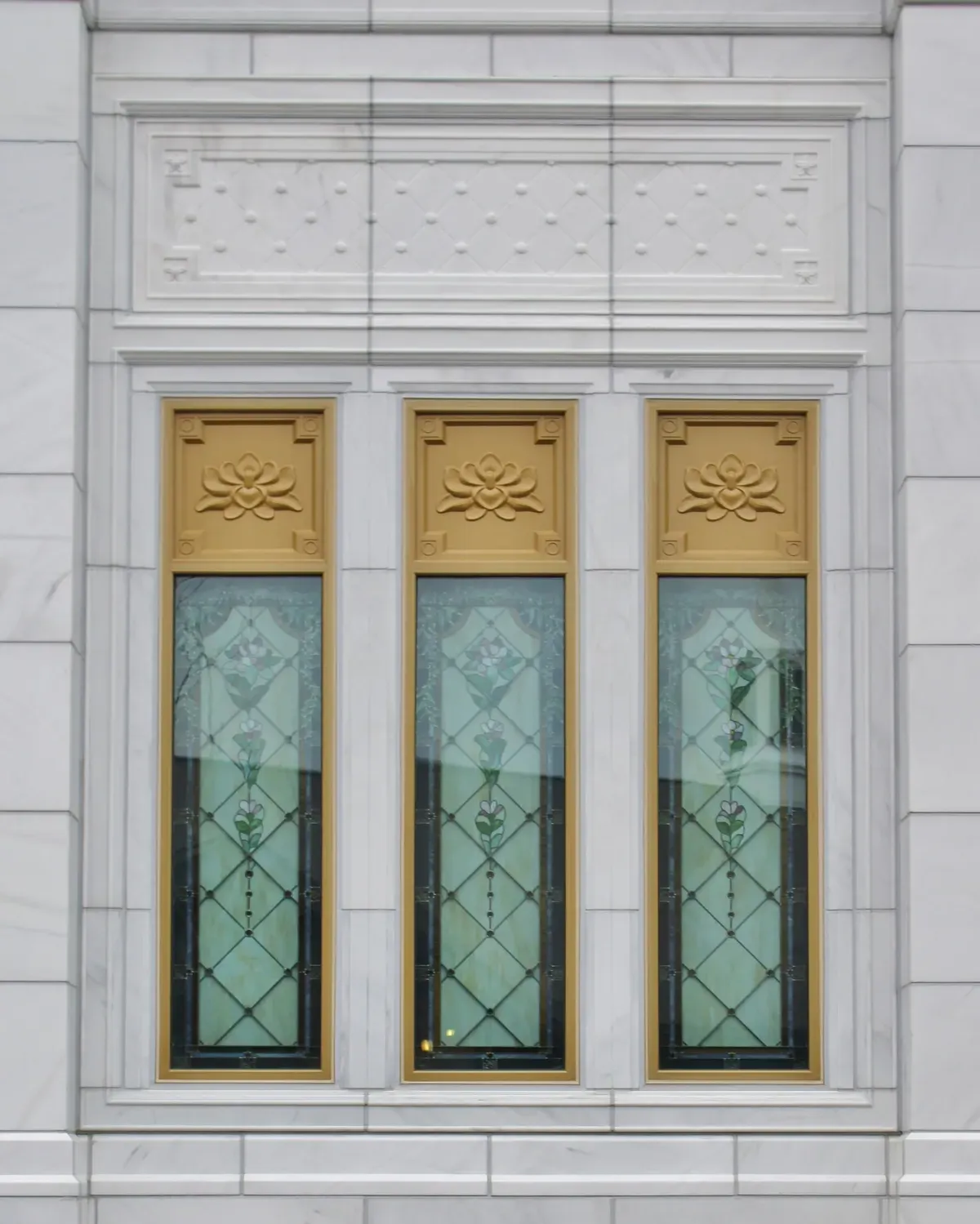 Three tall art glass windows with green and white floral patterns and gold frames carved with blossoms, set in white stone with a diamond lattice panel above.