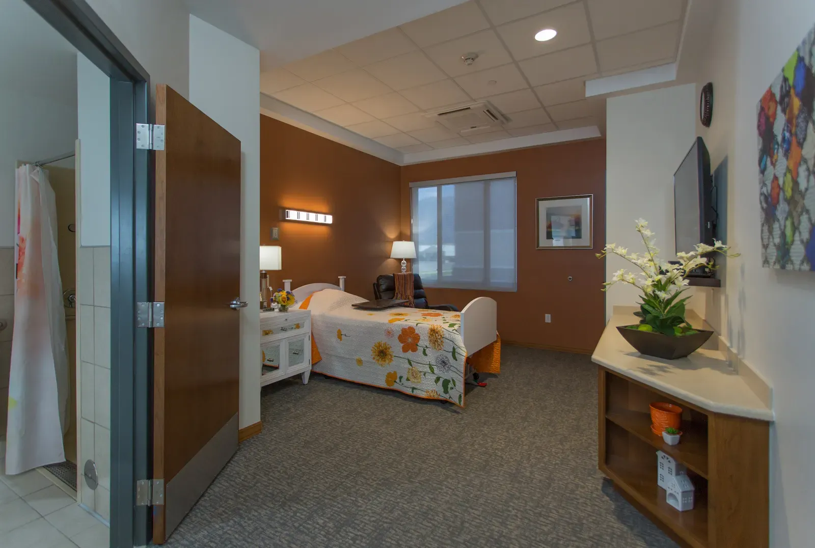 Resident room with a bed, a floral bedspread, a window, and a wall-mounted TV.