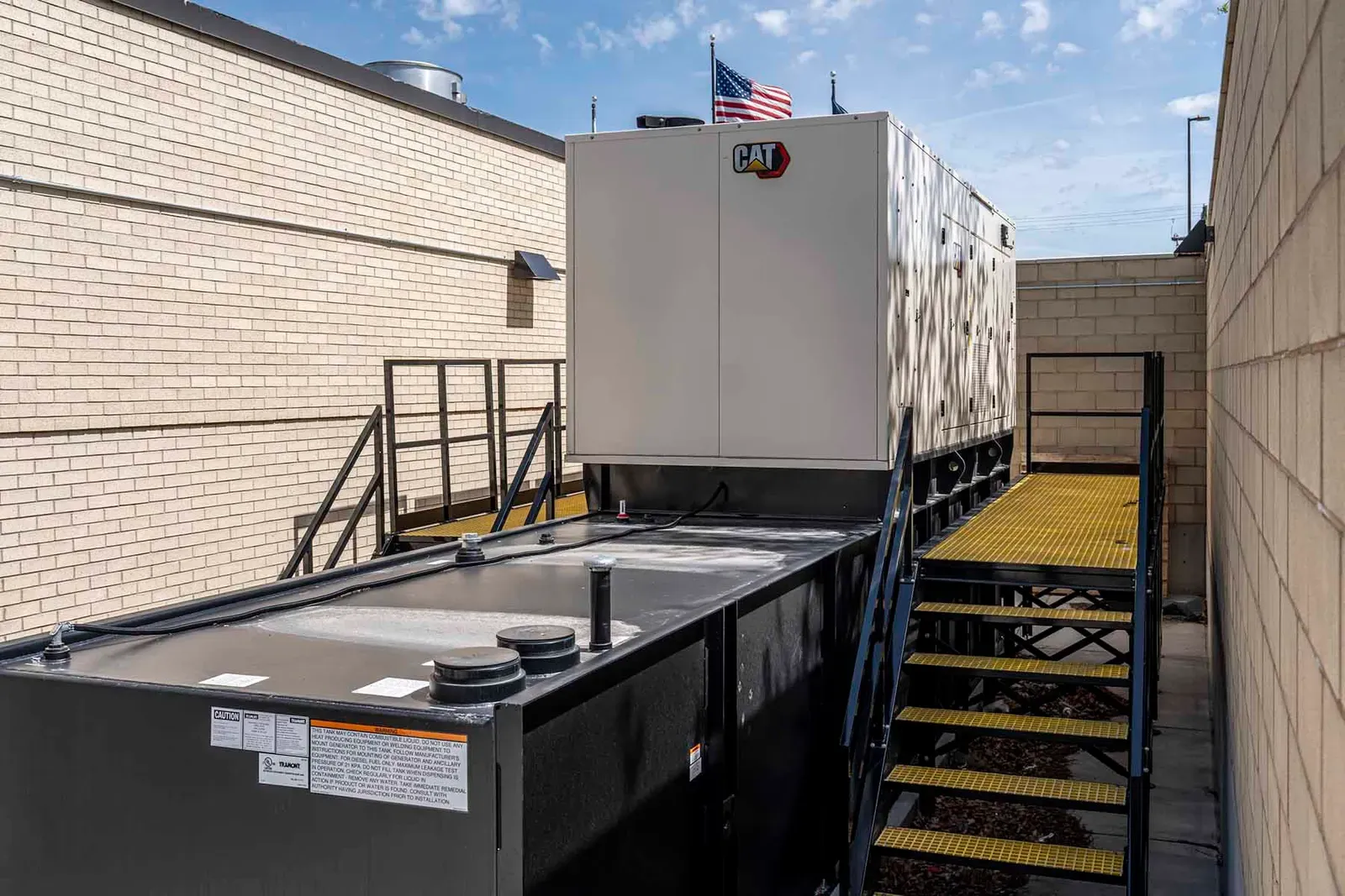Emergency generator in a white enclosure on a black steel base, with a steel stair and platform between brick walls.