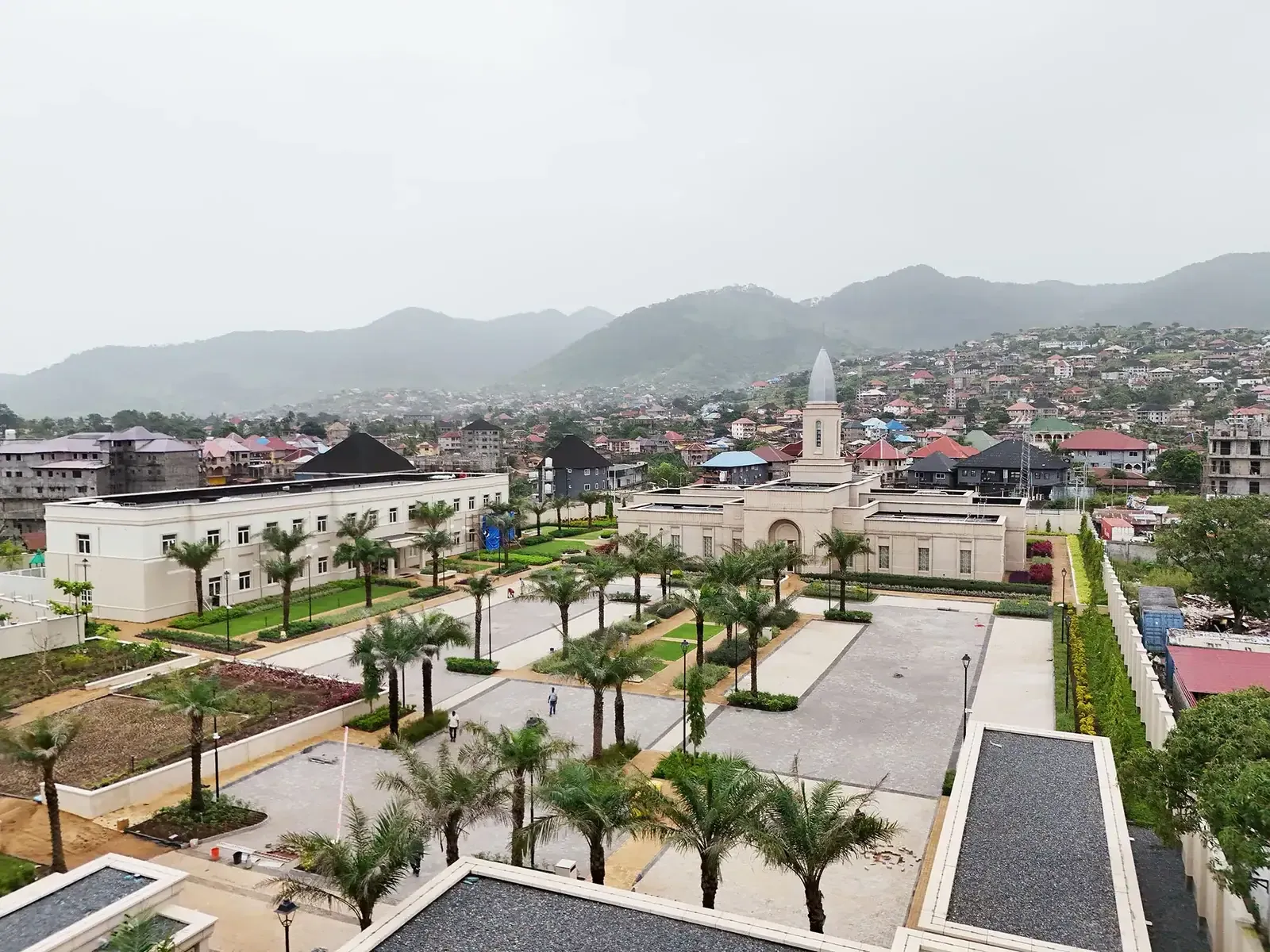 Aerial view of the temple campus with palm-lined courtyards and a long white building, with green hills and the city behind.