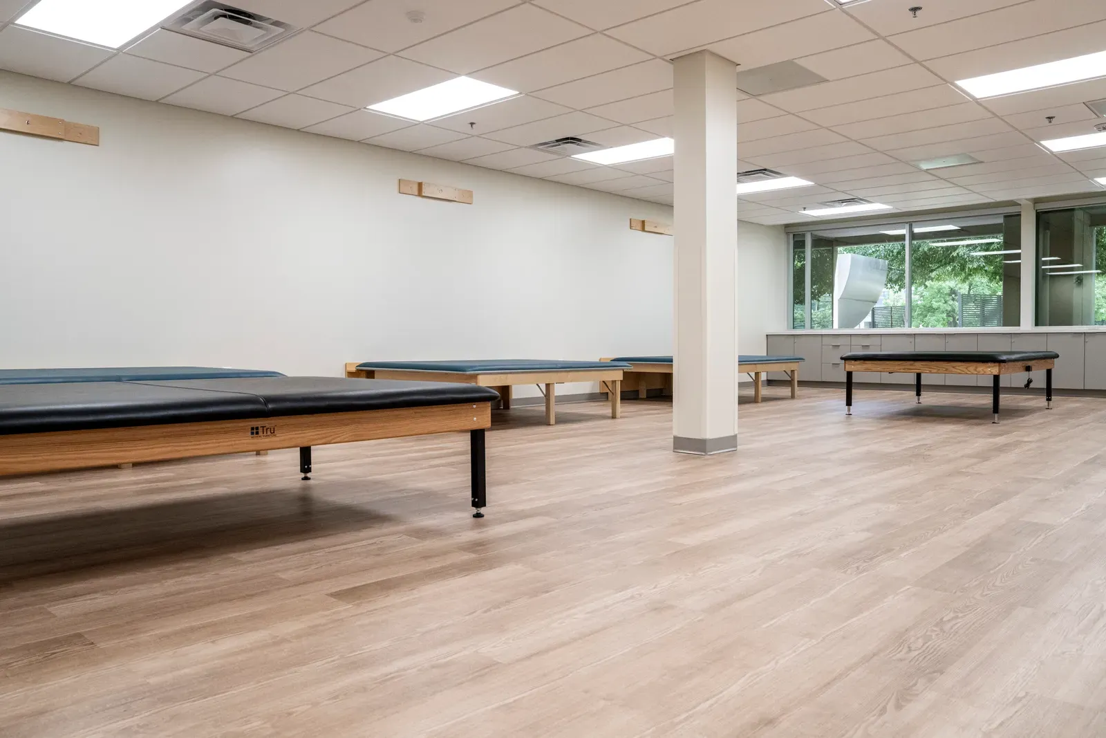 Therapy room with padded wooden treatment tables and a steel-trimmed column on a wood-look floor.