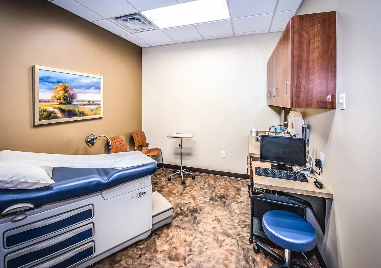 Exam room with an exam table, a desk and computer, and a landscape painting on the wall.
