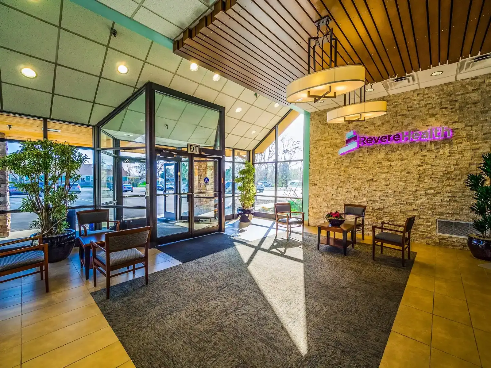 Lobby entrance with glass doors, a wood slat ceiling, a stone wall carrying the Revere Health sign and seating by the windows.