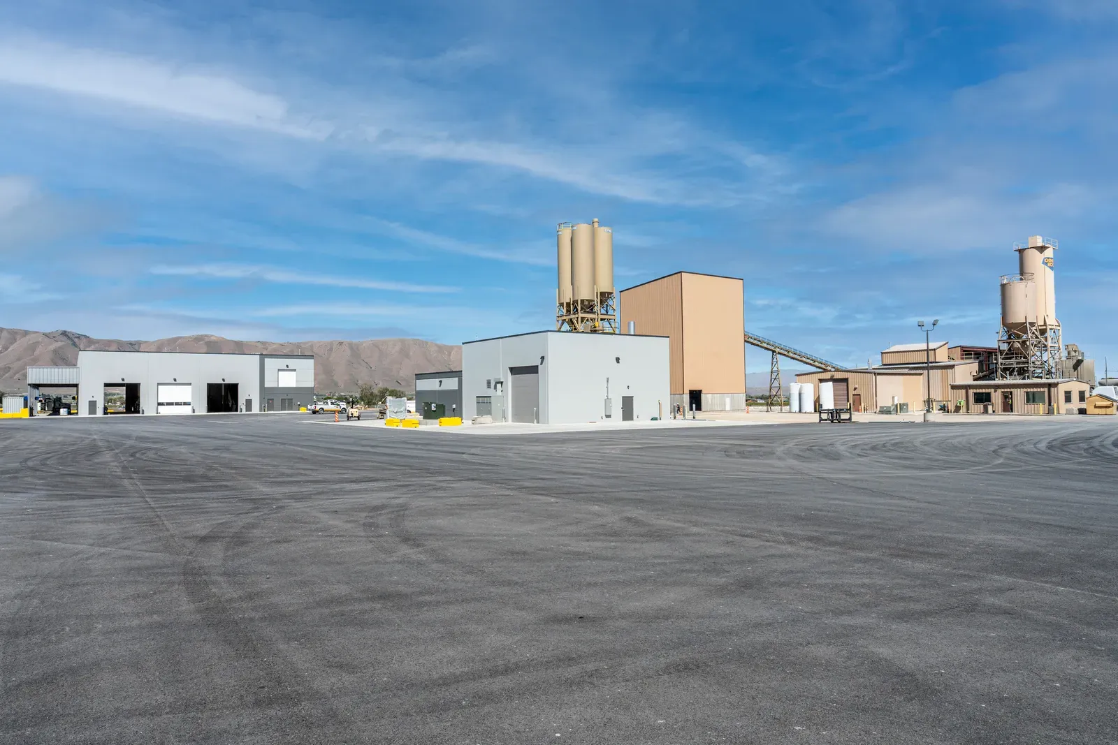 Wide view across the paved yard to gray buildings, tan silos and a conveyor with hills behind.