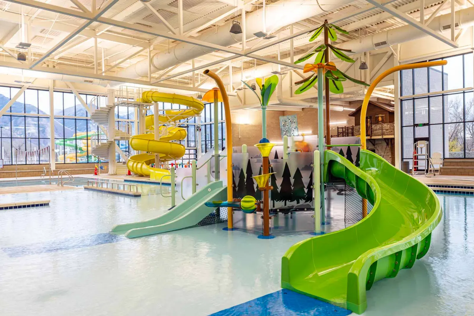 Indoor water play area with green and yellow slides and a tree-themed play structure, with tall windows looking out to the mountains.