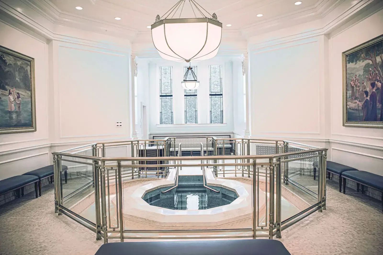 Baptismal font ringed by a glass and brass railing, with paintings on the walls and benches along the sides.