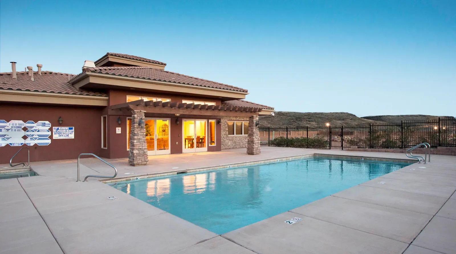 Swimming pool beside the lit clubhouse at dusk, with a stone-columned pergola and an iron fence.