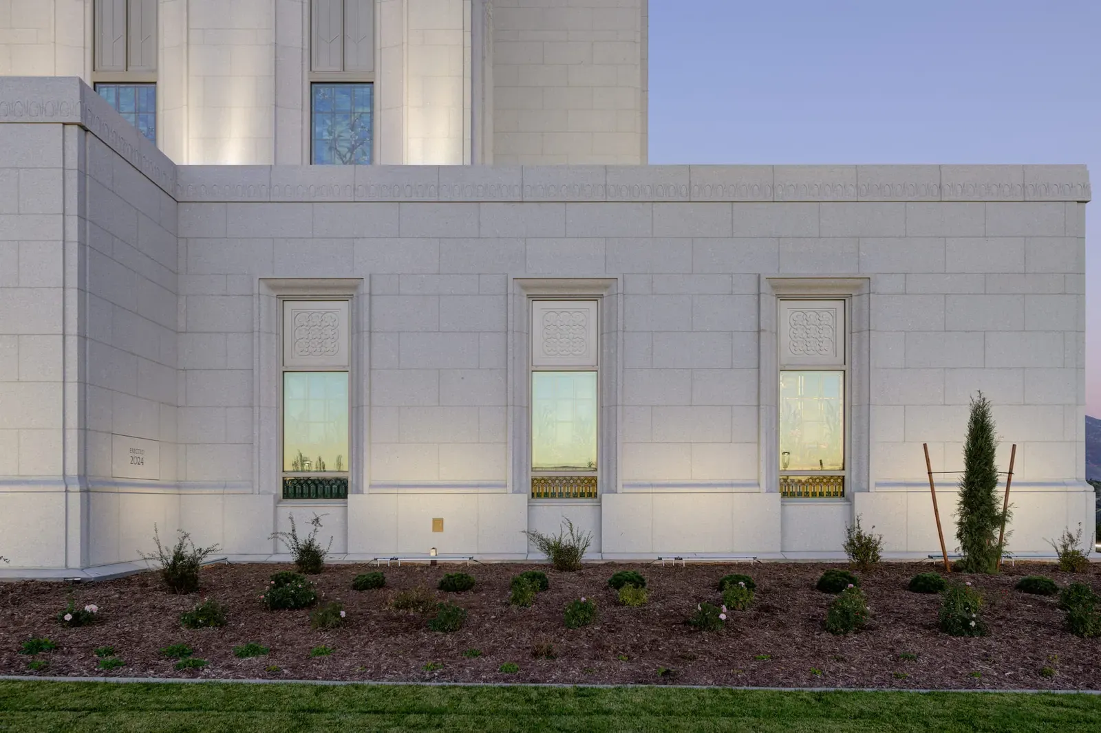 Stone side wall of the temple with three art glass windows and young landscaping.