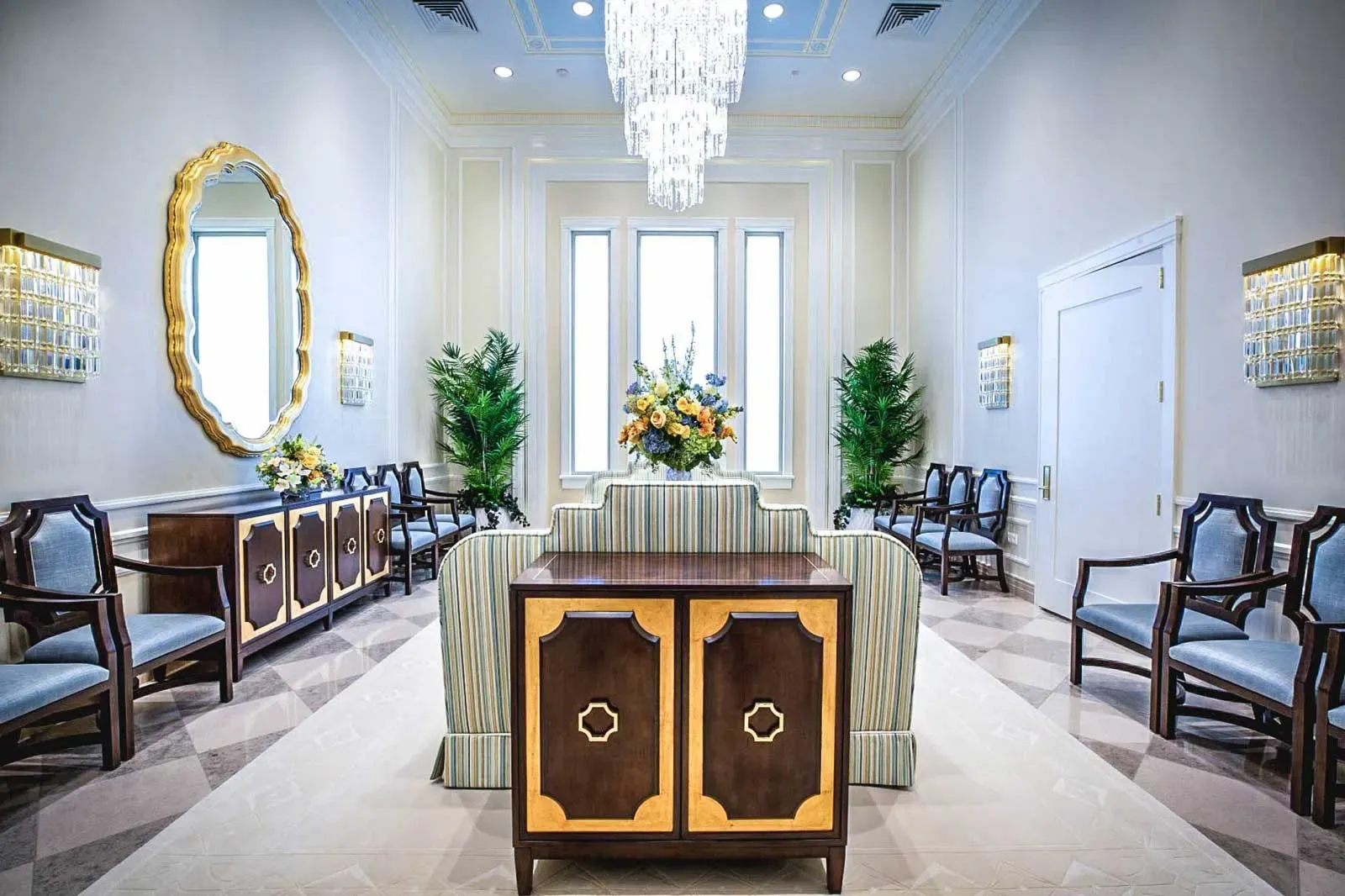 Sitting room with striped settees, a gold-framed oval mirror, a crystal chandelier and a bouquet of flowers.