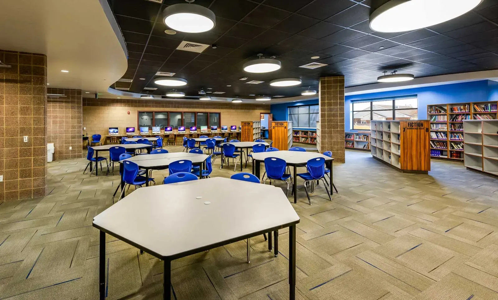 Library with white tables and blue chairs, tall wood shelving and a row of computers along the back wall