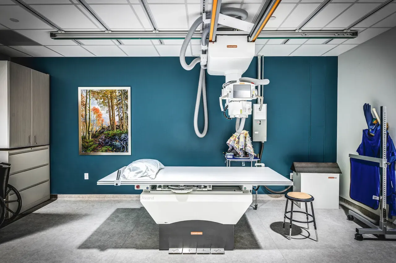 X-ray room with an overhead imaging unit, an exam table and a framed forest painting on a teal wall.