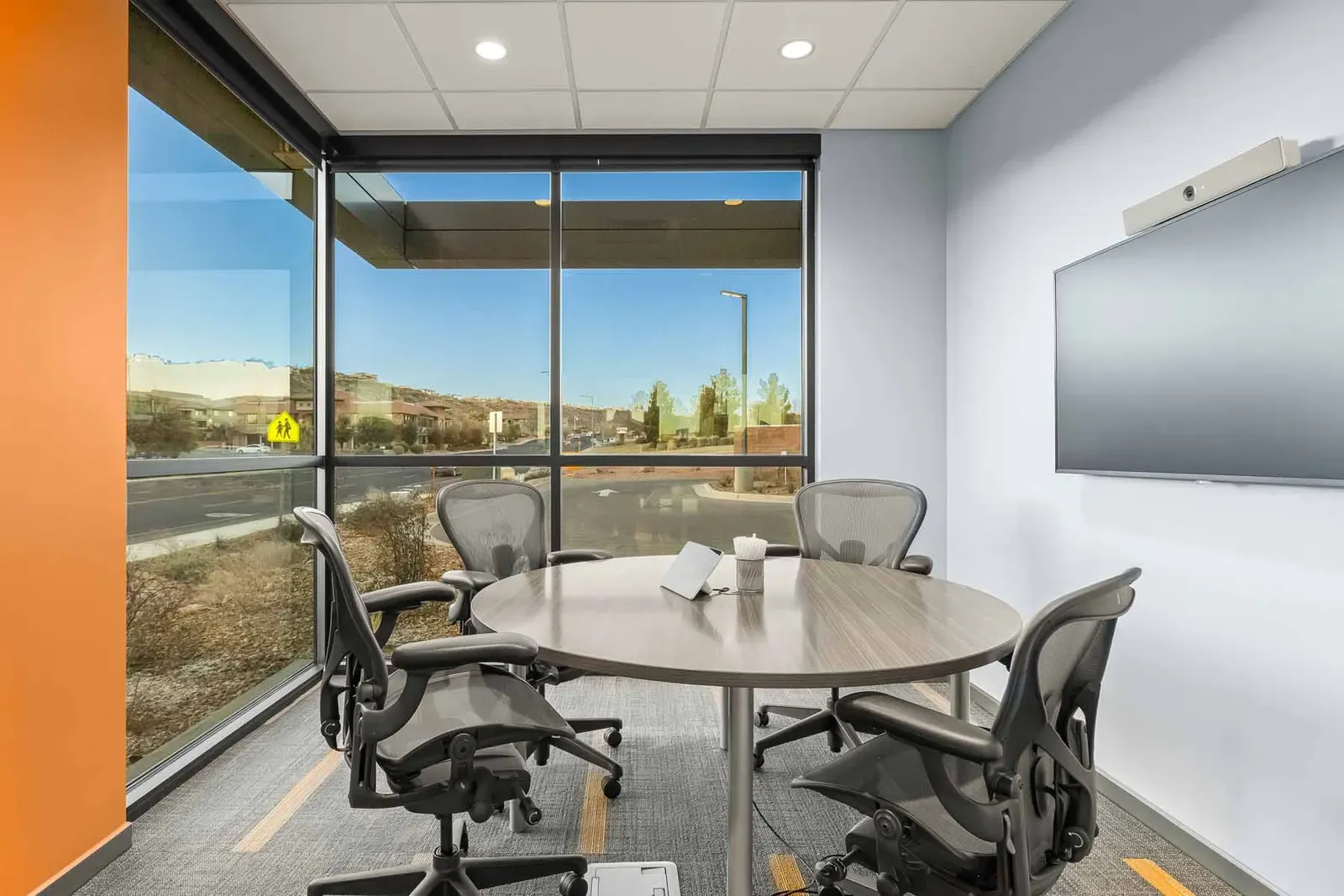 Small meeting room with a round table, four mesh chairs and a wall-mounted screen beside a corner window.