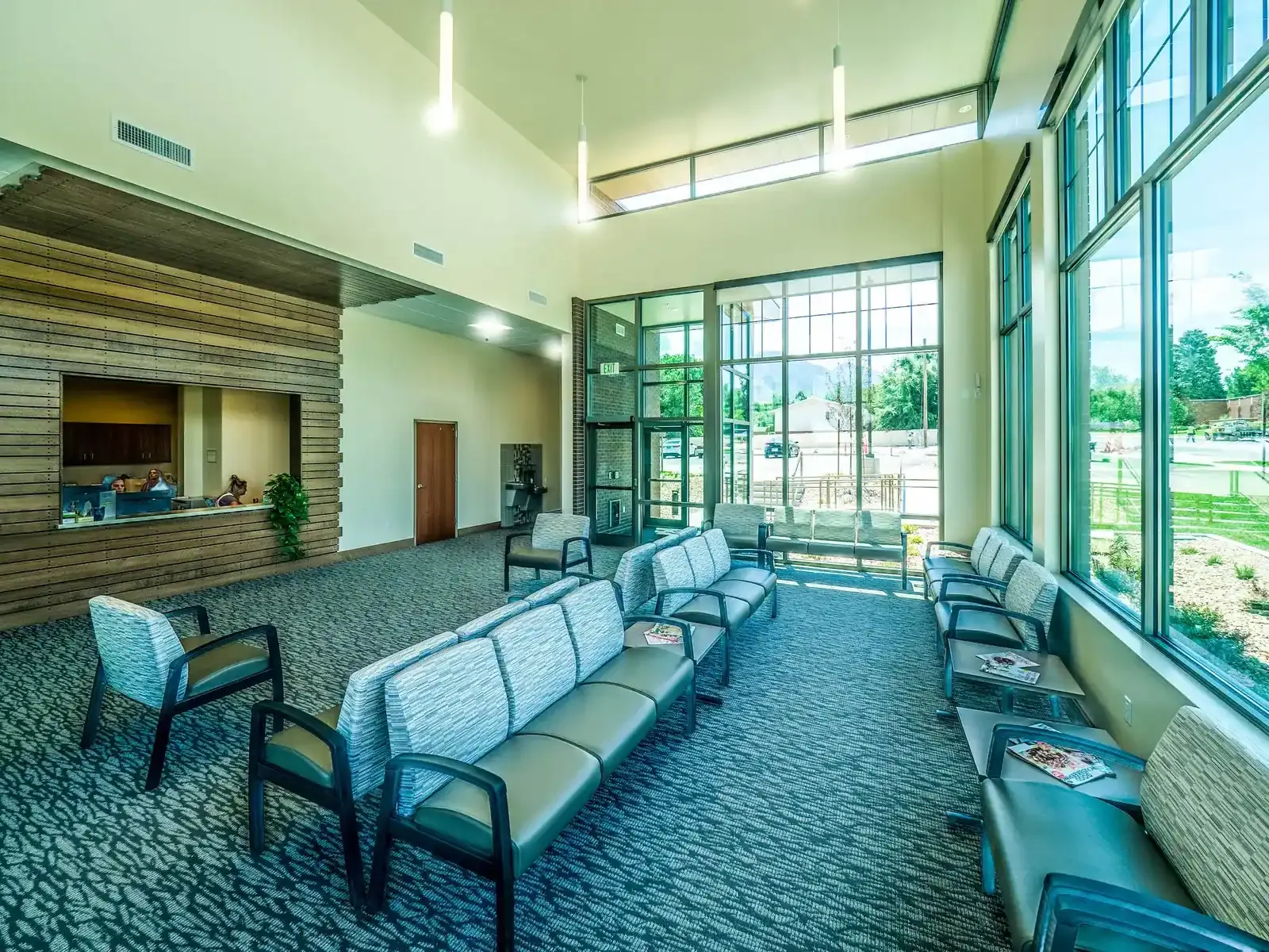 Waiting room with a wood-clad reception window, upholstered seating and a high ceiling with a wall of windows.