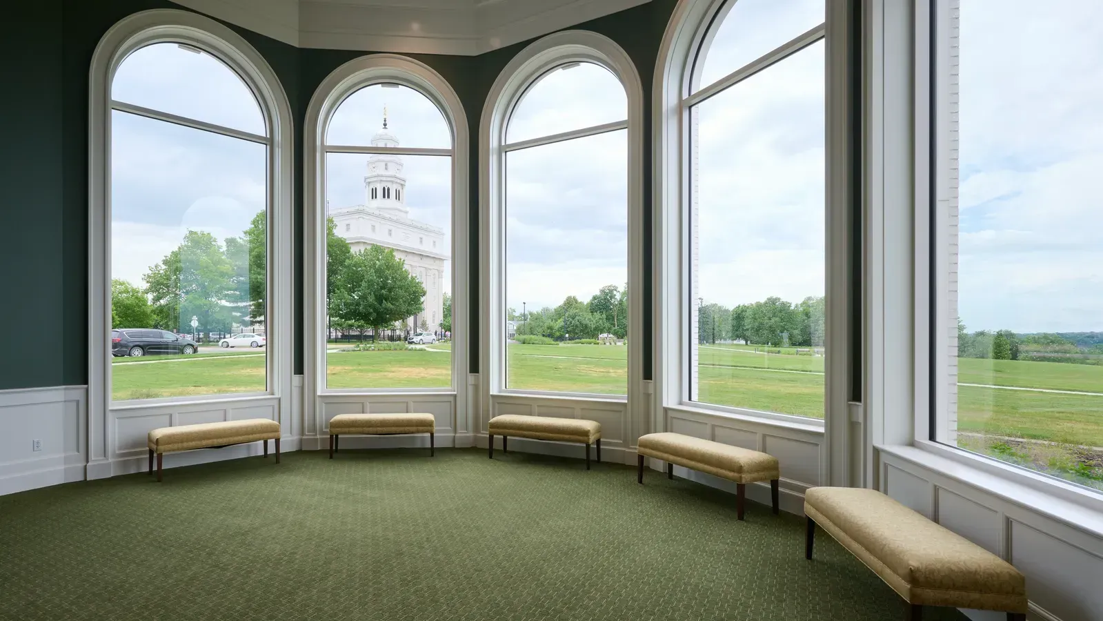 Room with a bank of tall arched windows and cushioned benches, framing a view of the white Nauvoo Illinois Temple across a green lawn.