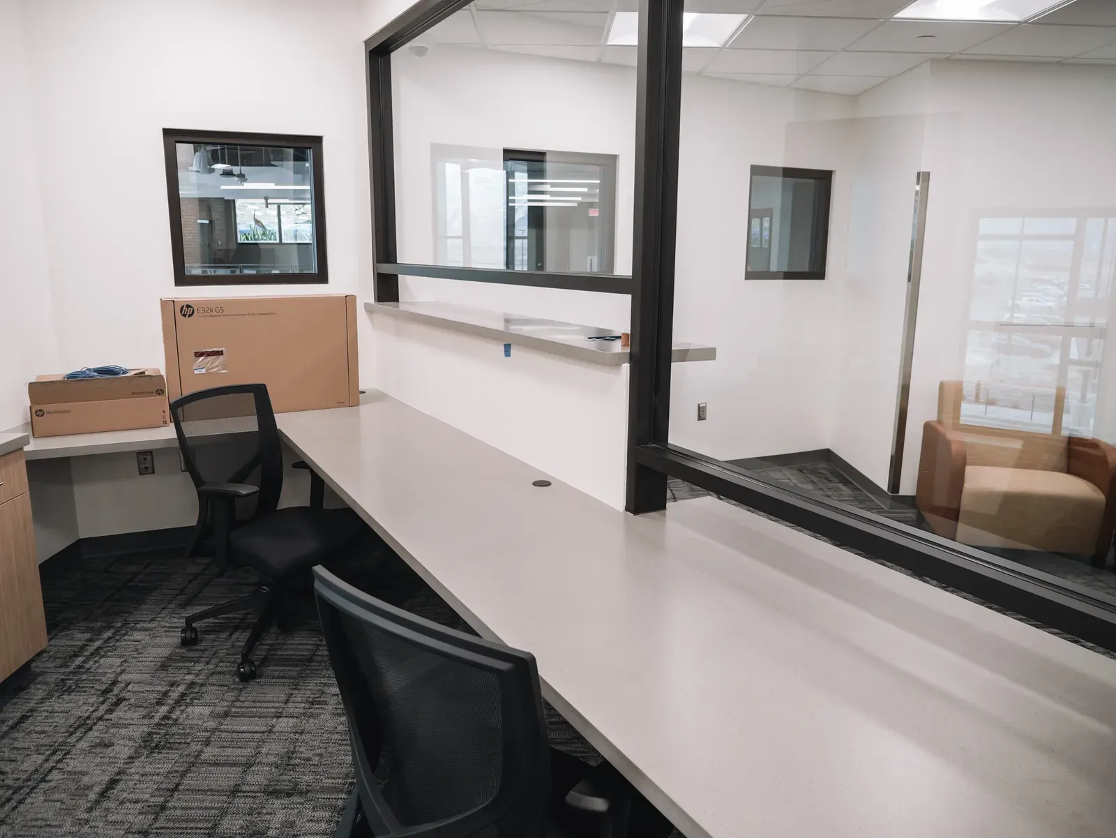 Office with a long work counter, black mesh chairs and a glass partition looking into a seating area.