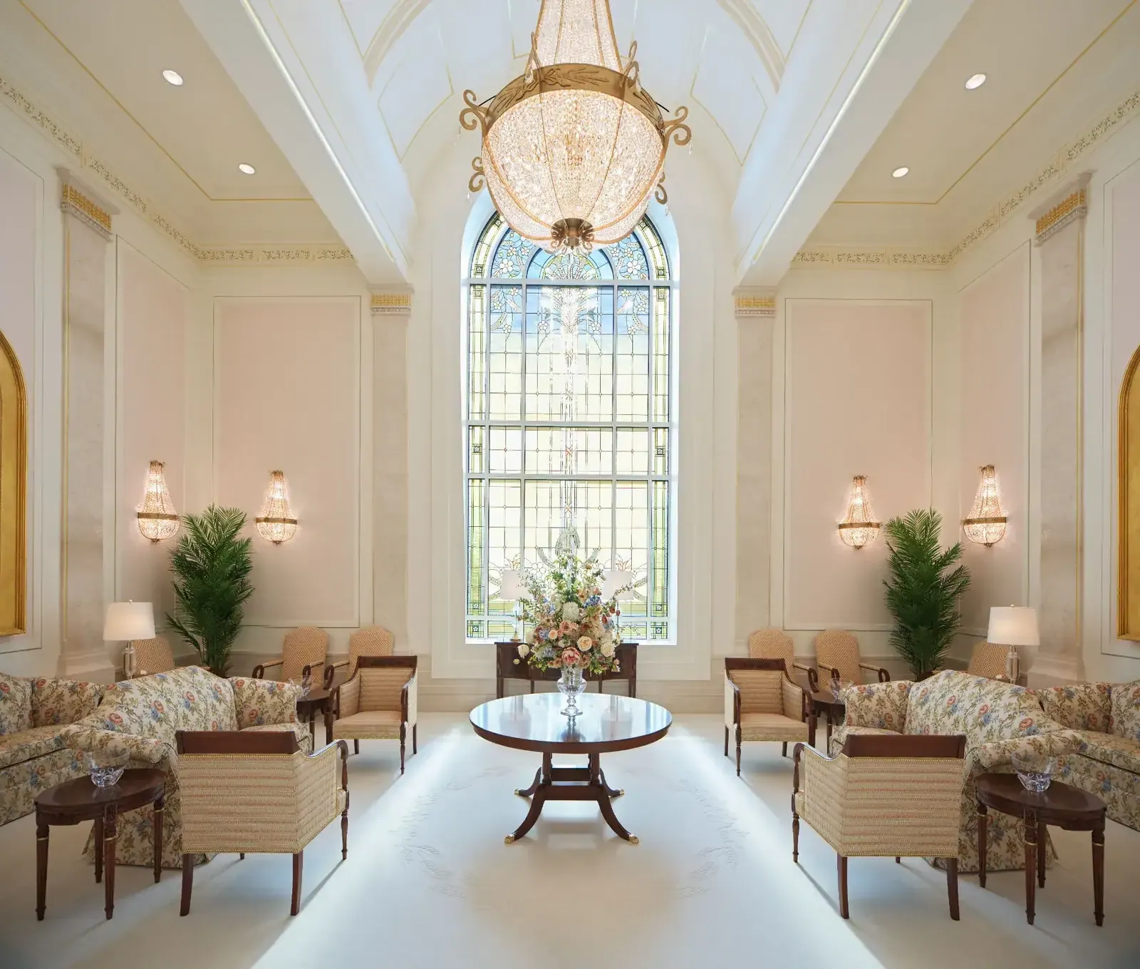 Room with a tall stained-glass window, a large crystal chandelier, floral sofas, armchairs and a round table with a flower arrangement