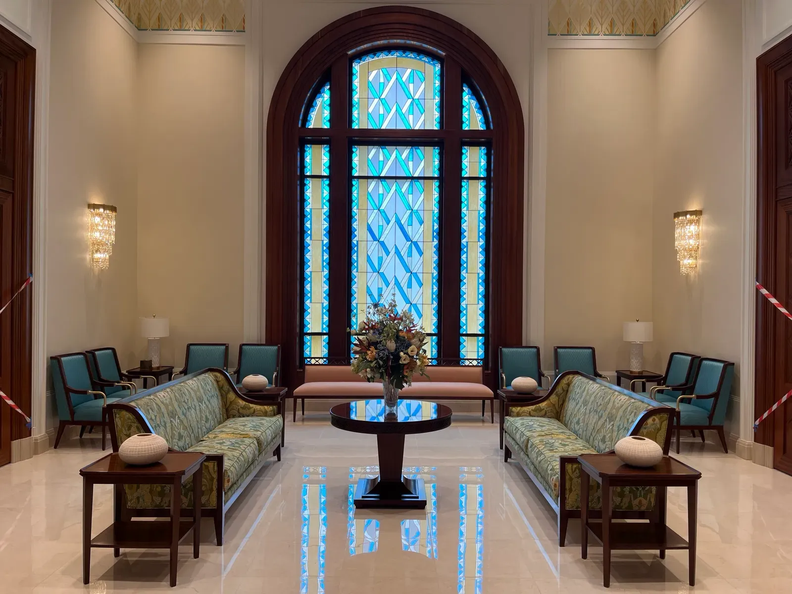 Sitting room with an arched blue and yellow art glass window, two patterned sofas, teal chairs and a round table with flowers.