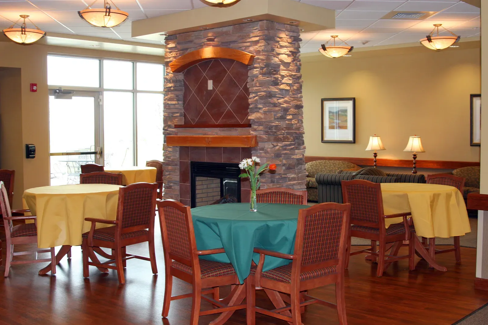 Dining area with round tables in yellow and teal cloths, wood chairs and a stone fireplace column.