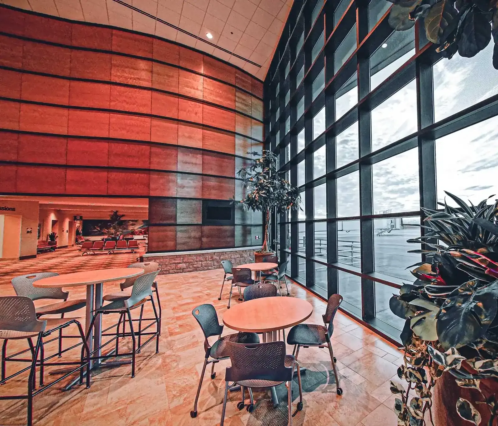 Two-story seating area with a red wood-paneled wall, a tall glass curtain wall facing the airfield and high bistro tables.