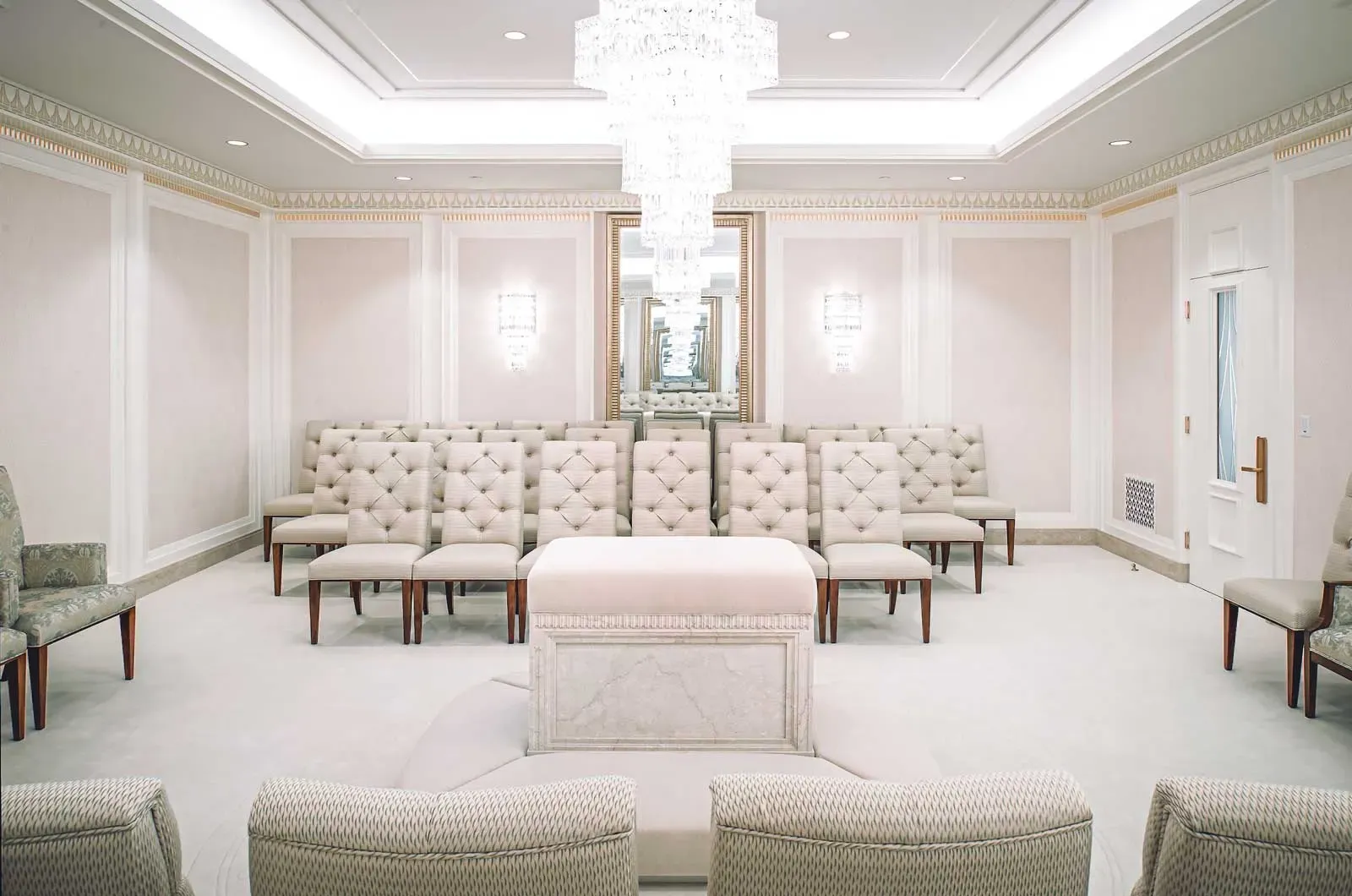 Quiet room with a central cushioned altar, rows of tufted chairs, and a crystal chandelier