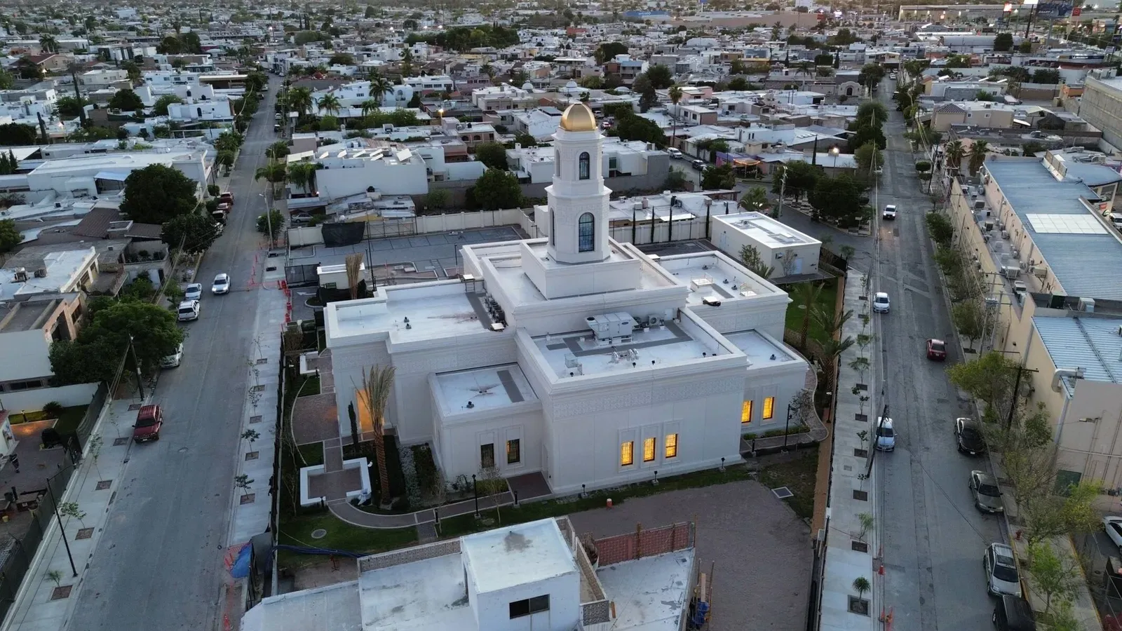 Aerial view at dusk of the temple among city blocks, with lit windows and the tower and gold dome rising above the roofs.
