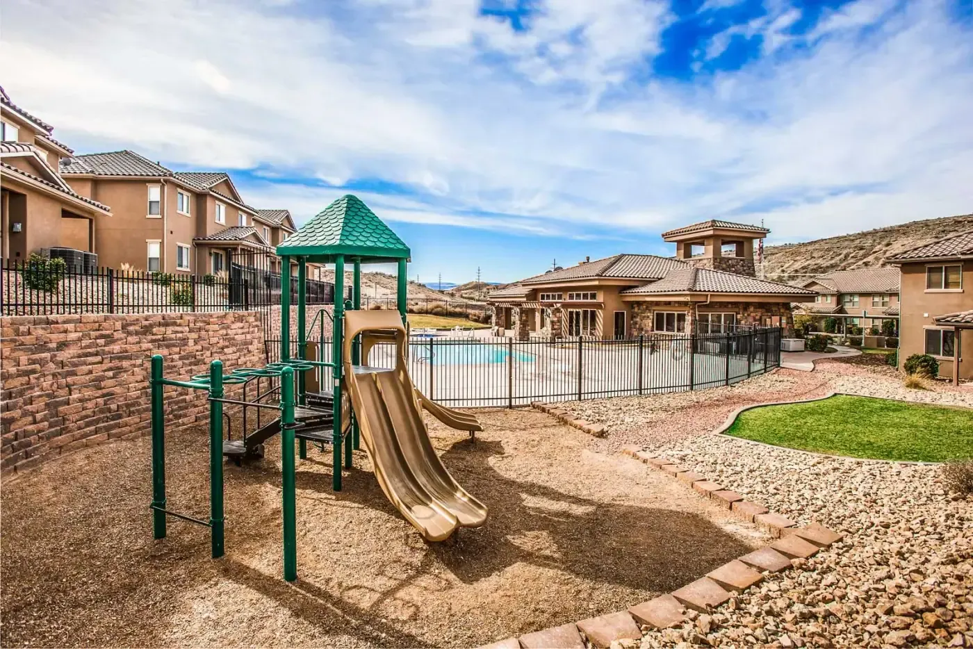 Playground with a green climbing structure and slide behind an iron fence, with the pool and clubhouse in the background