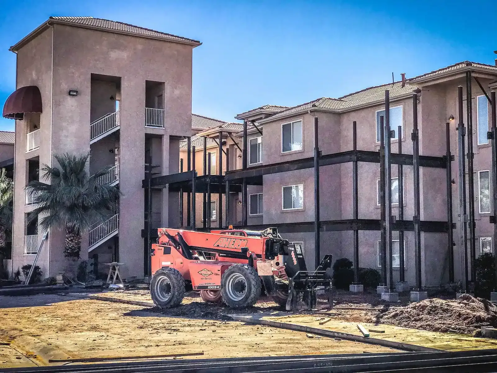 Stucco building under construction, with steel framing along its walkways and an orange telehandler in the foreground