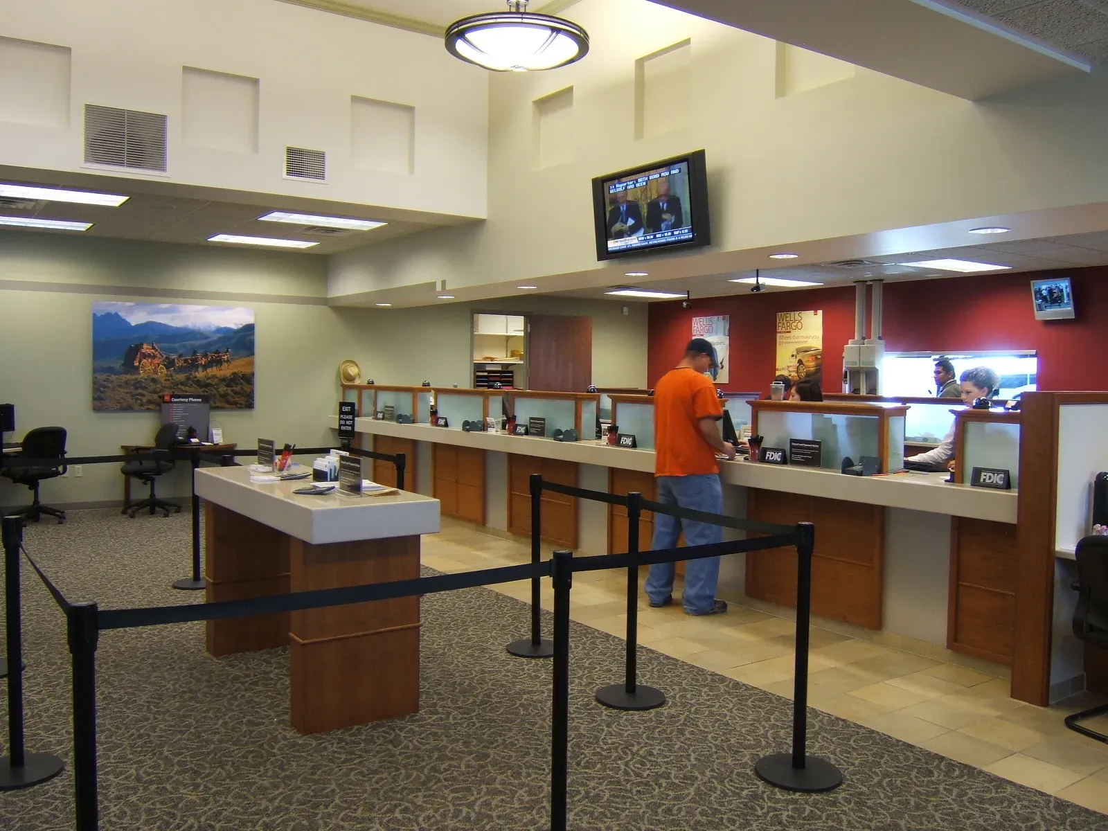 Banking lobby with a teller counter, queue stanchions and a mountain landscape painting on the wall.