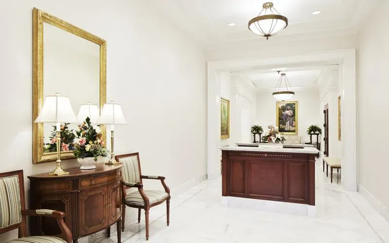Hall with a wood desk, a gold-framed mirror, a dresser with lamps and flowers, and a framed painting beyond