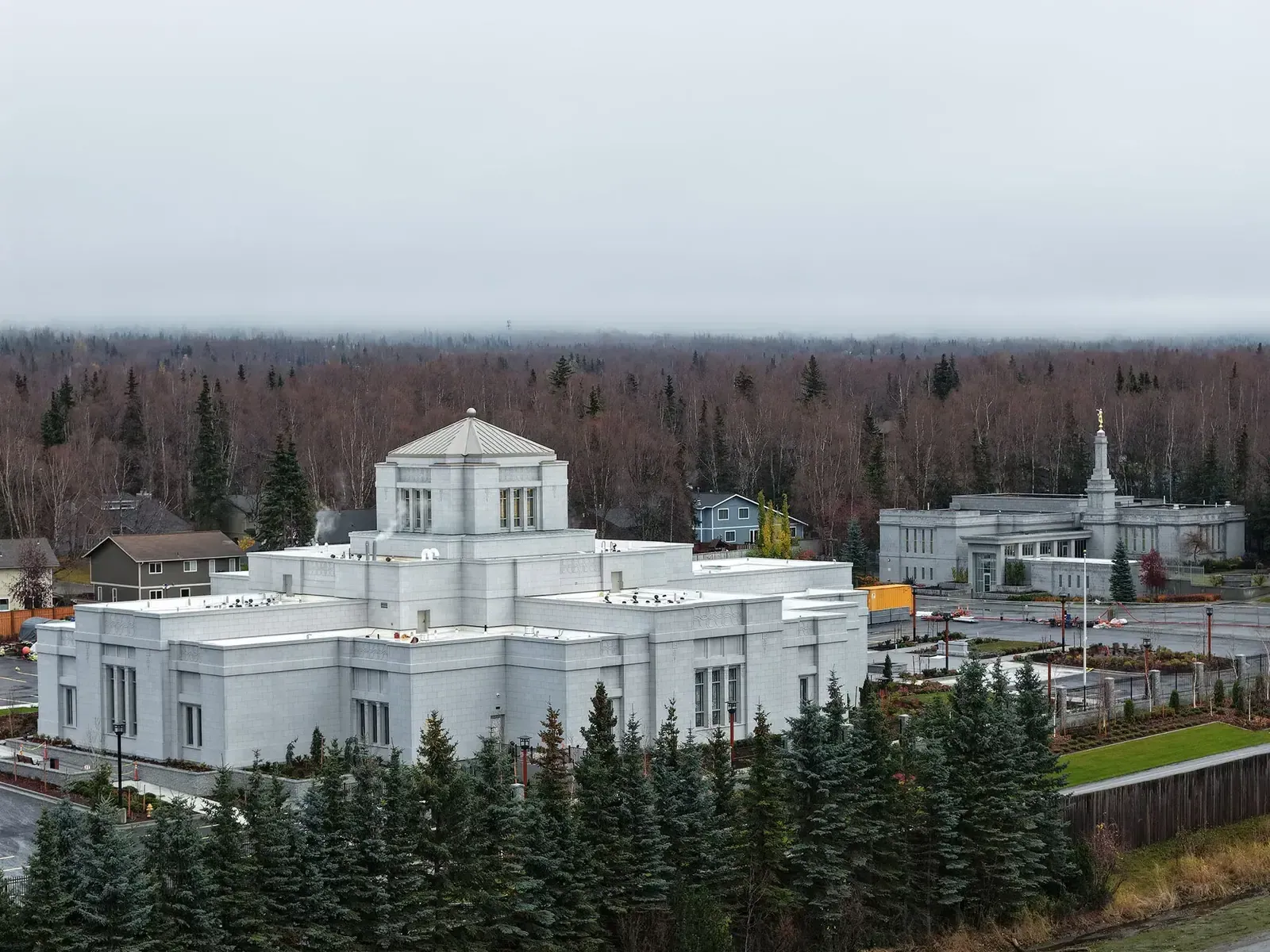 Aerial view of the new temple among spruce trees, with a smaller building topped by a spire at the right.